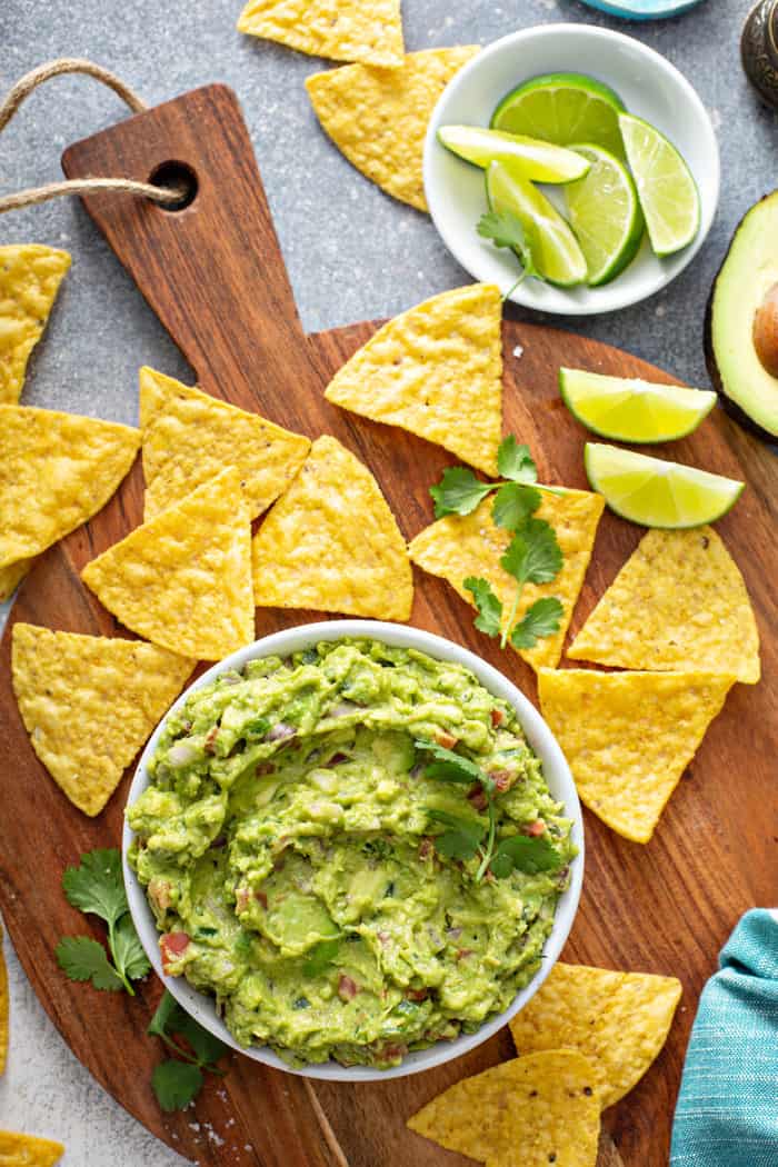 Overhead view of a bowl of spicy guacamole on a wooden board, surrounded by tortilla chips and slices of lime