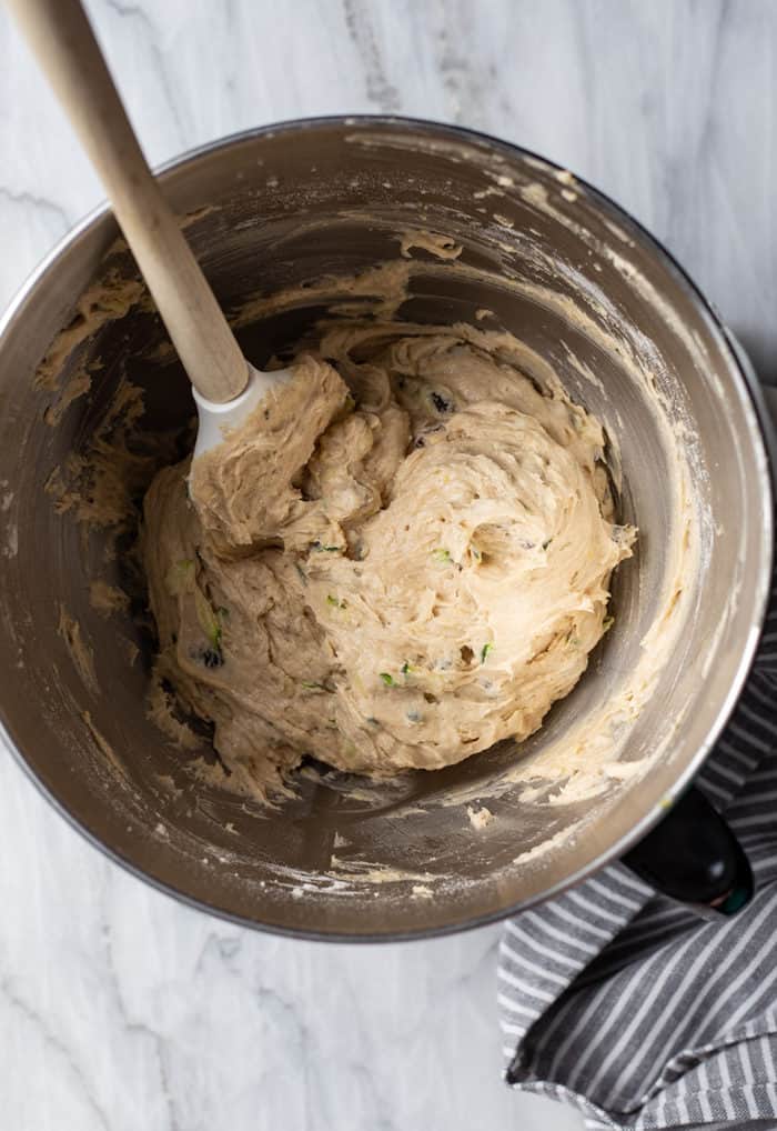 Spatula stirring together zucchini cookie dough in a metal mixing bowl on a marble countertop