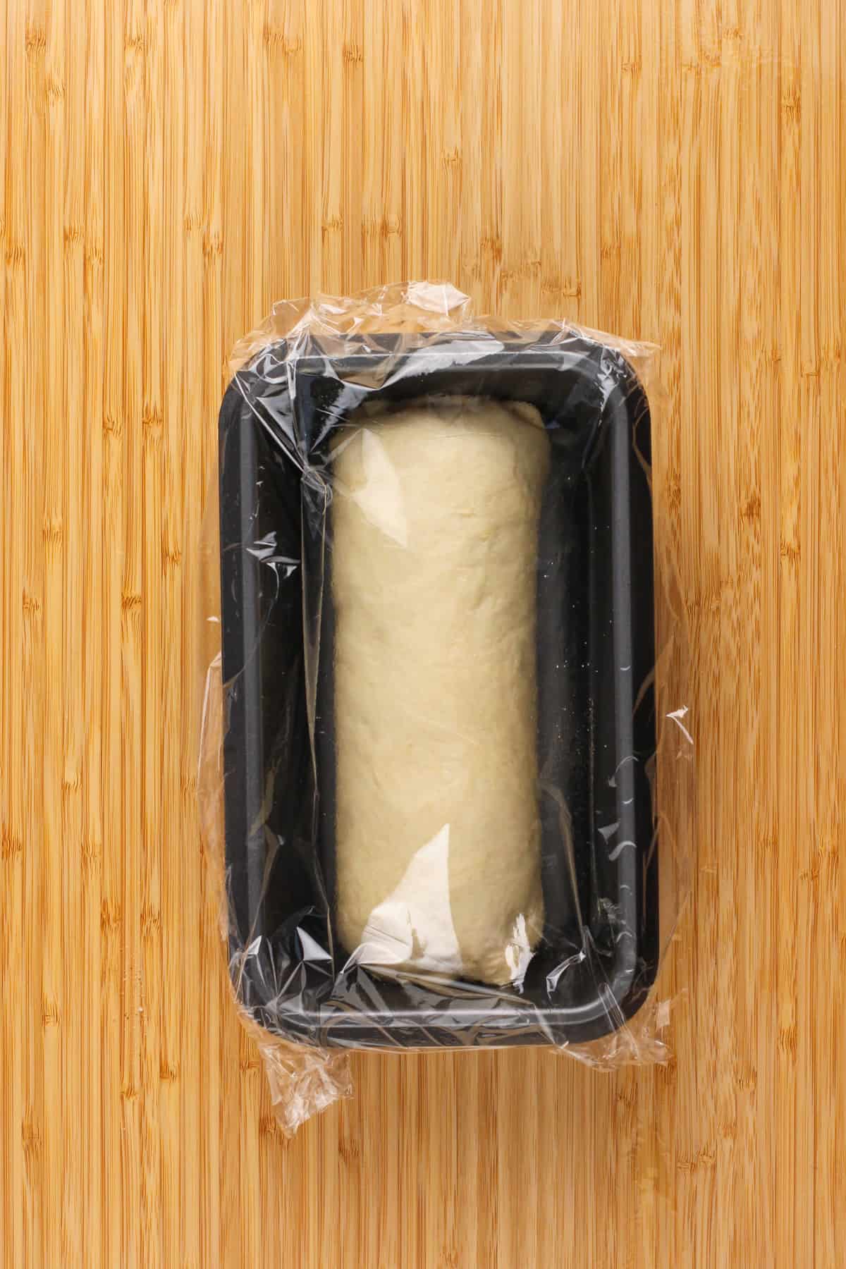 Assembled loaf of cinnamon swirl bread in a loaf pan covered with plastic wrap, ready for proofing.