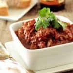 Bowl of prime rib chili next to a spoon on a wooden surface