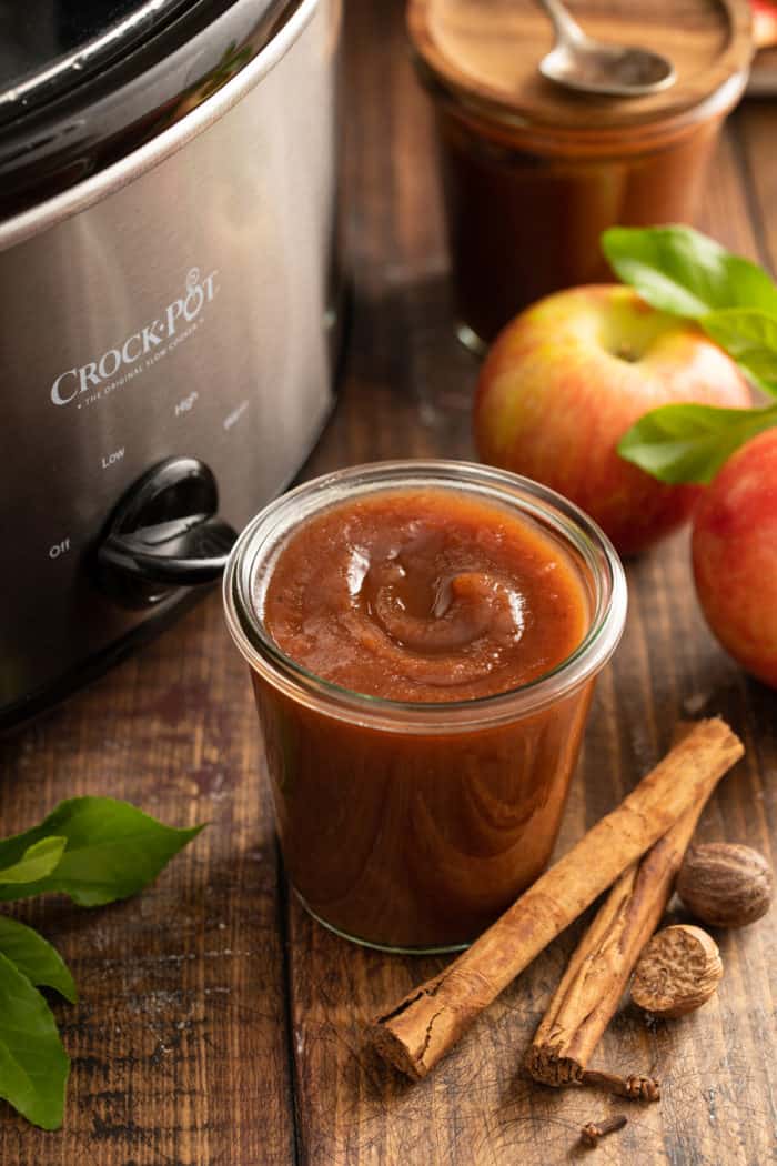 Open glass jar of apple butter set next to a slow cooker on a wooden tabletop.