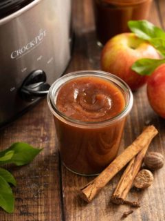 Open glass jar of apple butter set next to a slow cooker on a wooden tabletop.