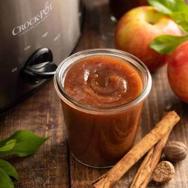 Open glass jar of apple butter set next to a slow cooker on a wooden tabletop.