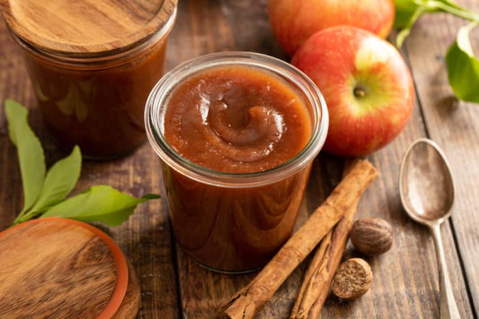 Open glass jar of apple butter set on a wooden tabletop, surrounded by whole apples and spices.