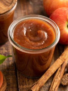 Glass jar filled with apple butter, surrounded by apples and whole spices.
