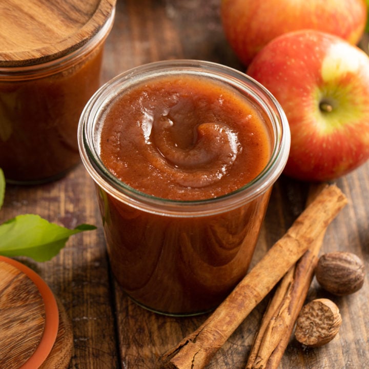 Glass jar filled with apple butter, surrounded by apples and whole spices.