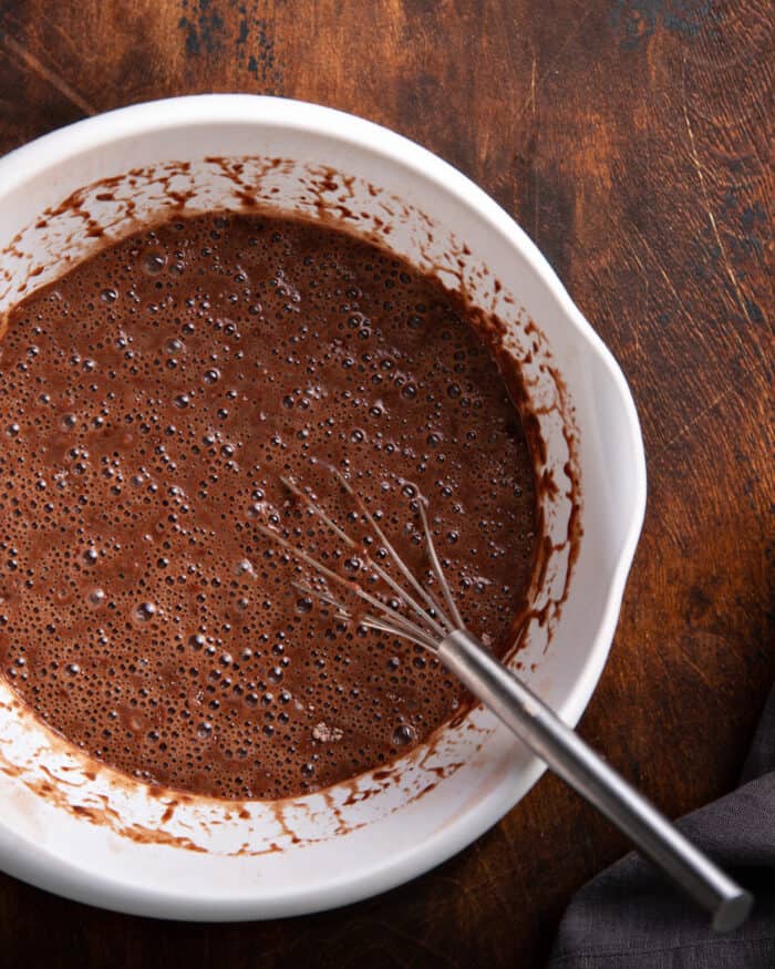 Chocolate cake batter and a whisk in a white mixing bowl on a wooden surface