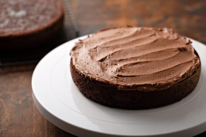 Single layer of chocolate cake topped with chocolate frosting on a white cake stand on a wooden background