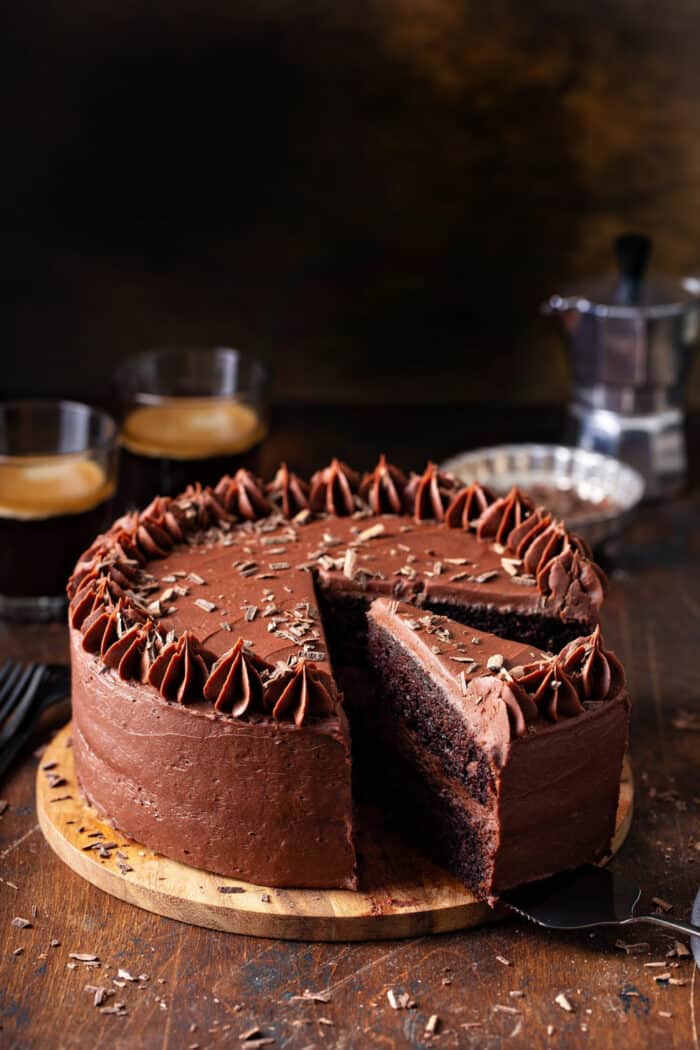 Chocolate cake covered in chocolate frosting on a wooden cake plate with a slice being removed