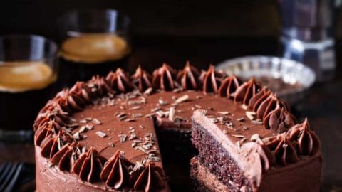 Slice being removed from a double layer chocolate cake with chocolate frosting, with coffee in the background