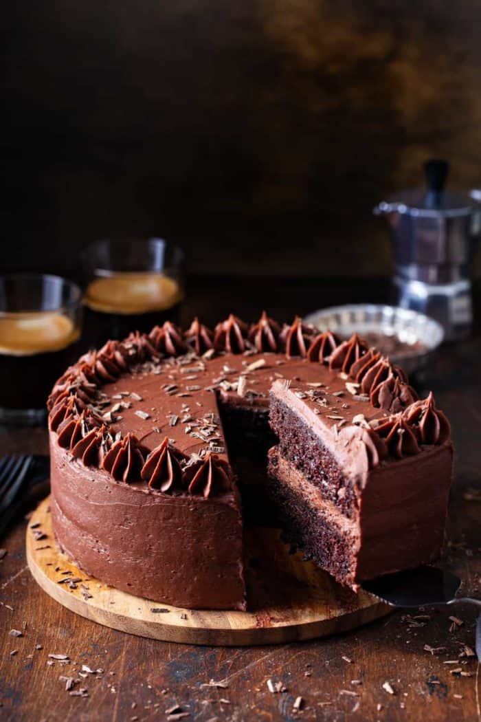 Slice being removed from a double layer chocolate cake with chocolate frosting, with coffee in the background