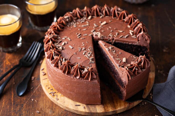 3/4 view of chocolate cake with chocolate frosting on a wooden cake stand with a slice being removed