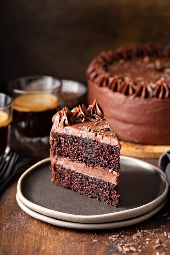 Slice of chocolate cake with chocolate frosting on a gray plate with coffee and chocolate cake in the background