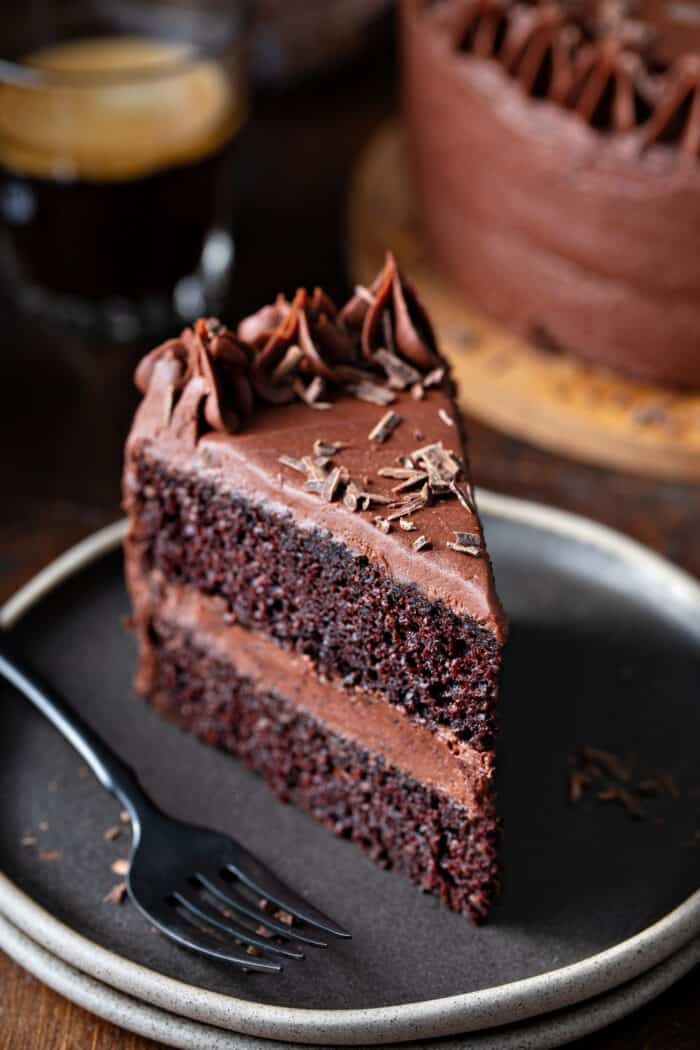 Fork next to a slice of chocolate cake with chocolate frosting on a gray plate, with coffee and chocolate cake in the background