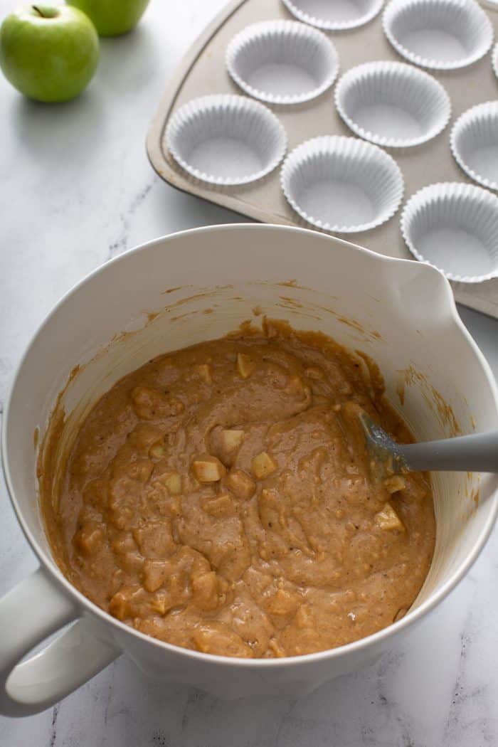 Apple cinnamon muffin batter and a gray spatula in a white mixing bowl with a muffin tin lined with paper liners in the background