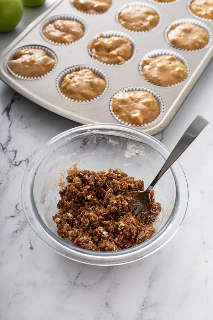 Streusel topping for apple cinnamon muffins in a glass mixing bowl with a muffin tin filled with muffin batter in the background