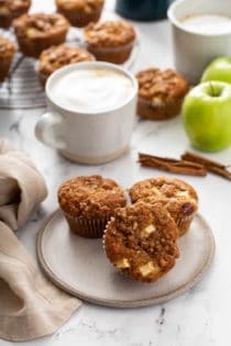 Three apple cinnamon muffins on a cream plate with lattes and more muffins on a cooling rack in the background