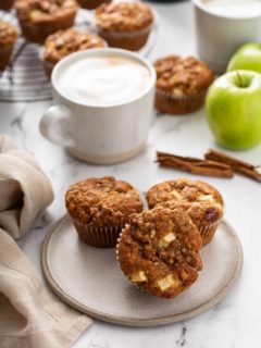 Three apple cinnamon muffins on a cream plate with lattes and more muffins on a cooling rack in the background