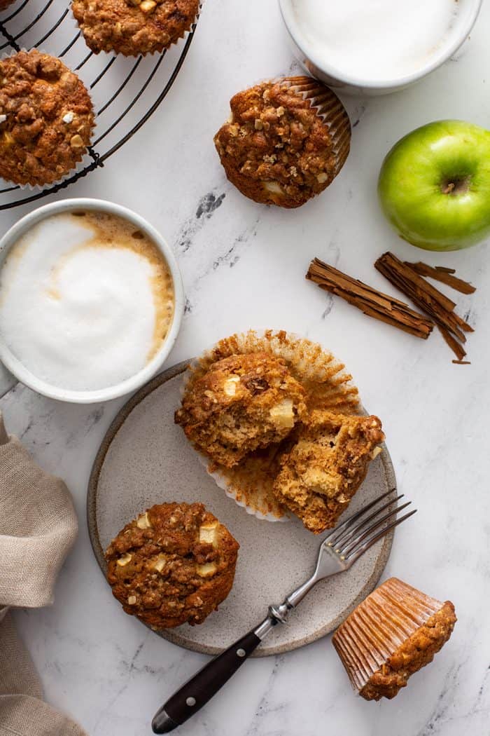 Overhead view of two apple cinnamon muffins next to a fork on a gray plate, surrounded by lattes, apples and more muffins