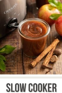 Open glass jar of apple butter set on a wooden tabletop, surrounded by whole apples and spices. Text overlay includes recipe name.