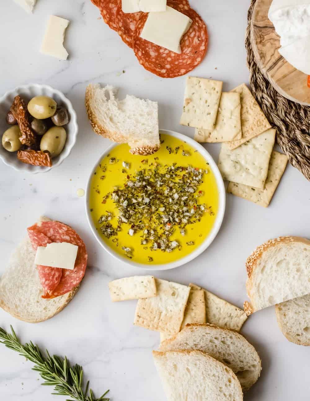 Shallow bowl of olive oil herb dip on a white platter with bread, olives and cured meats