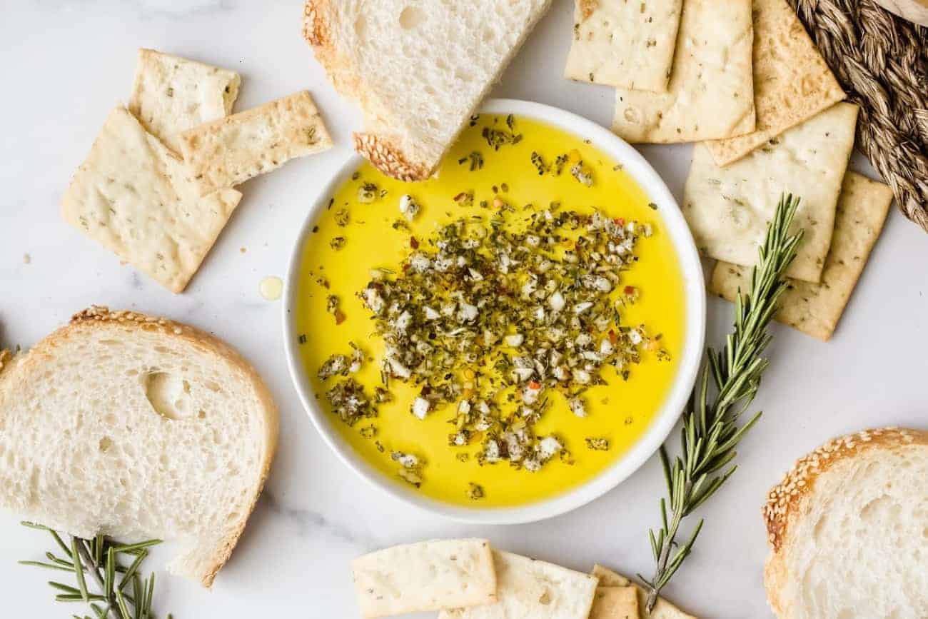 Bread and crackers surrounding a white bowl of olive oil herb dip