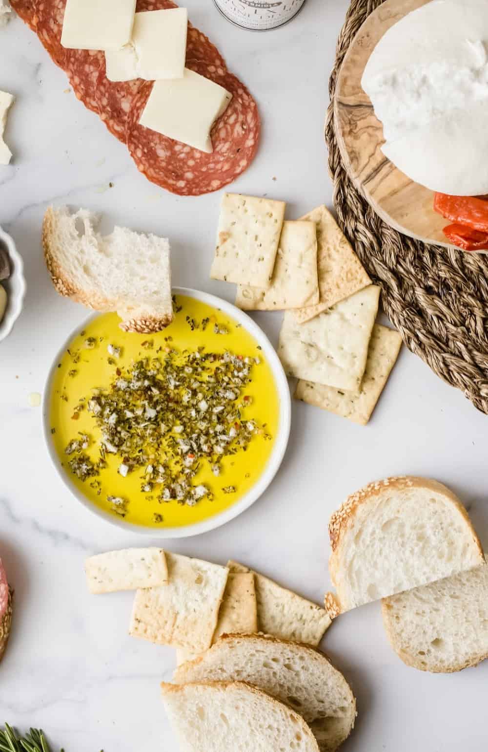 White bowl of olive oil herb dip on a white surface surrounded by bread and charcuterie