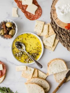 Spoon in a bowl of olive oil herb dip surrounded by bread, olives and charcuterie