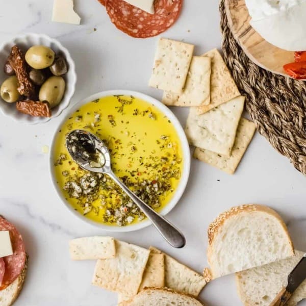 Spoon in a bowl of olive oil herb dip surrounded by bread, olives and charcuterie