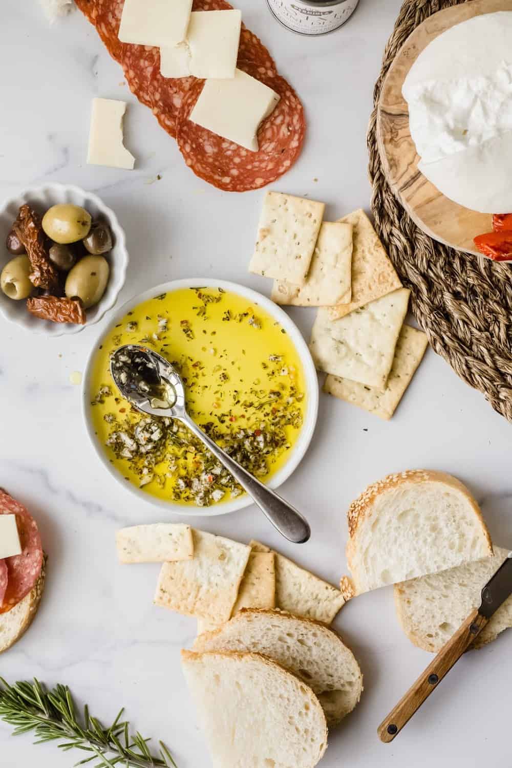 Spoon in a bowl of olive oil herb dip surrounded by bread, olives and charcuterie