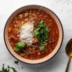 Bowl of sausage and lentil soup on a white surface, garnished with parsley and shredded parmesan