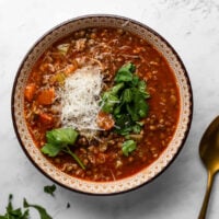 Bowl of sausage and lentil soup on a white surface, garnished with parsley and shredded parmesan