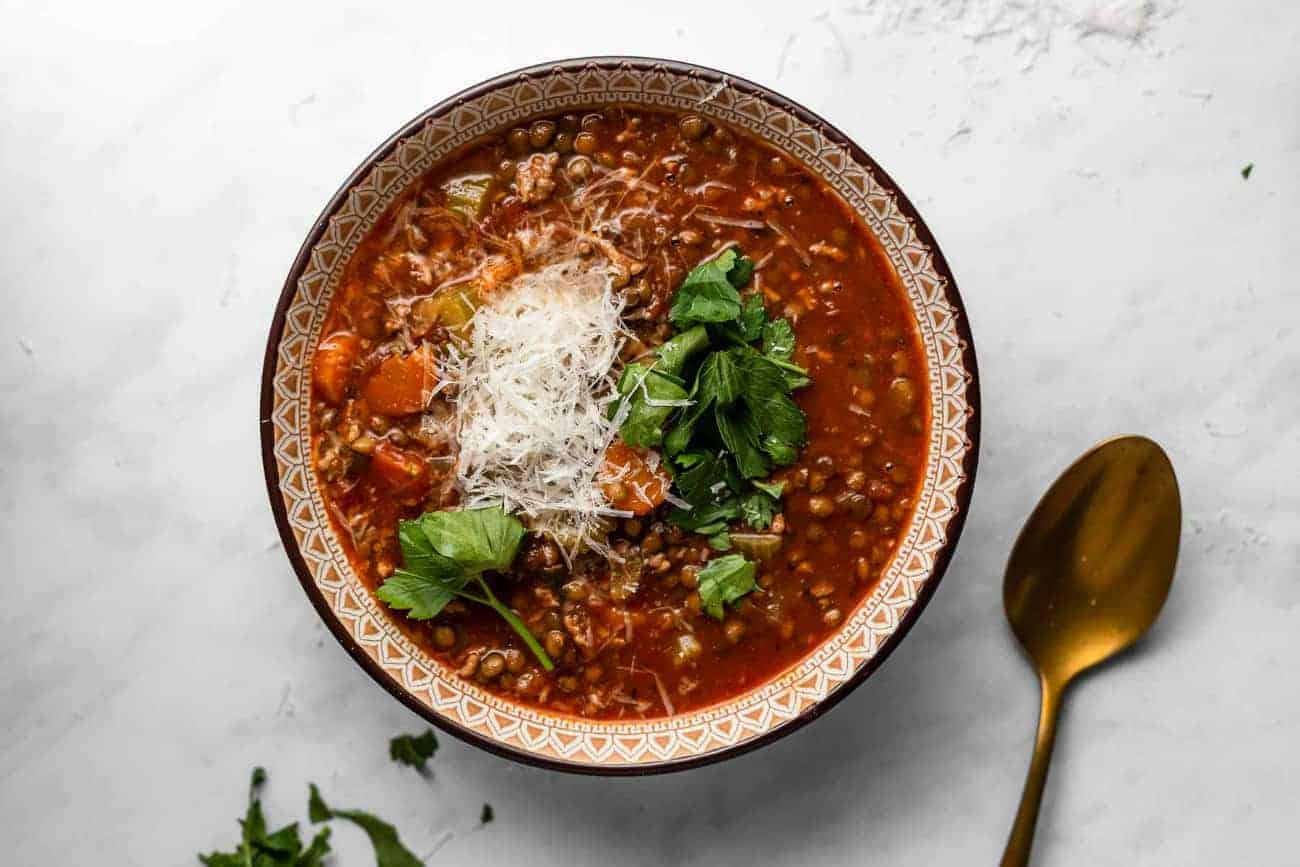 Bowl of sausage and lentil soup on a white surface, garnished with parsley and shredded parmesan