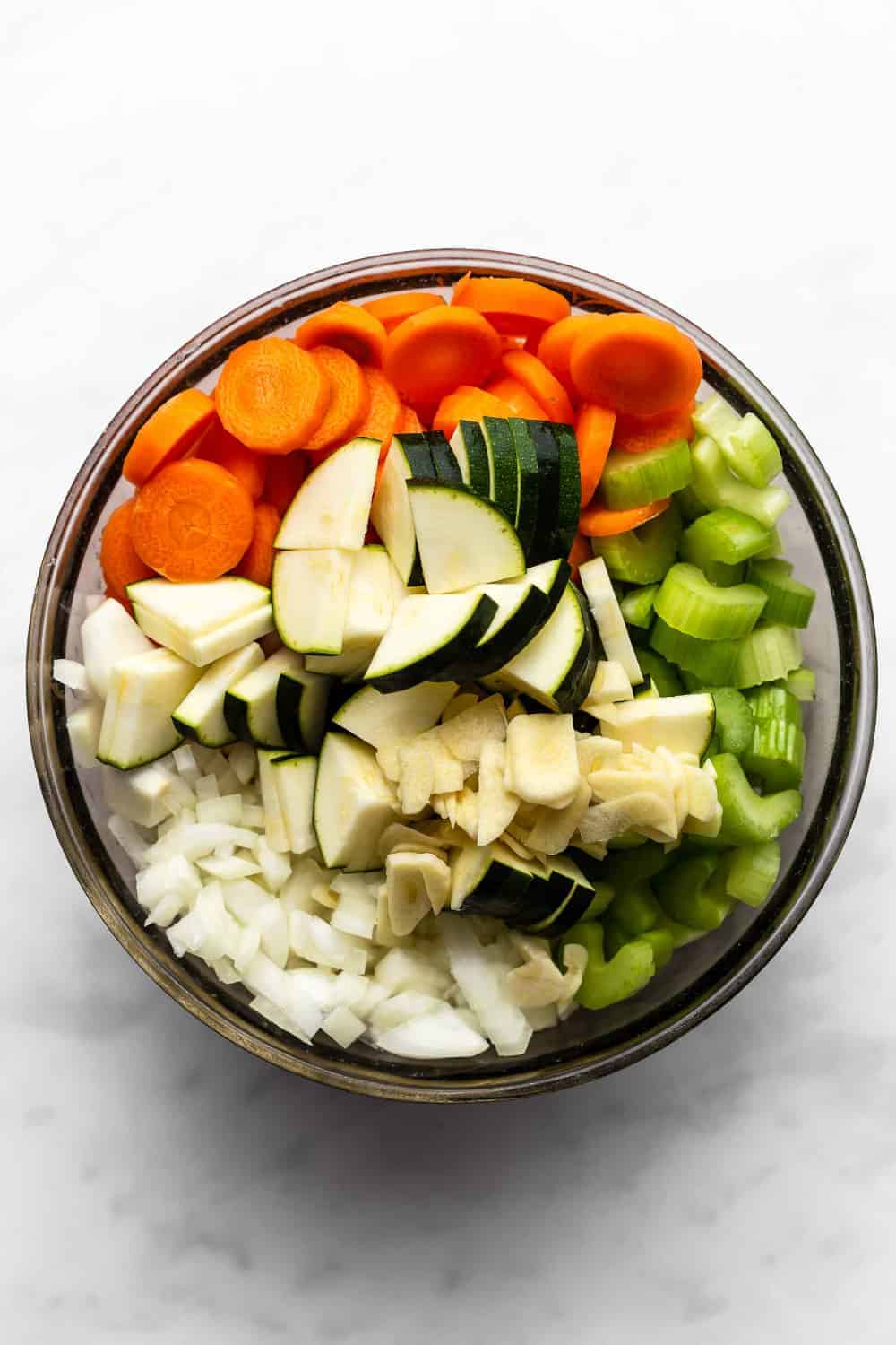 Overhead shot of diced onions, carrots, celery and zucchini in a bowl on a white surface