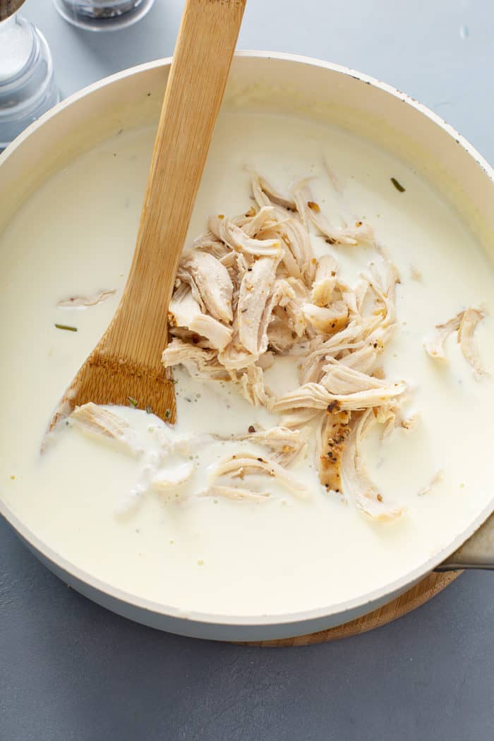 Wooden spoon stirring shredded roast chicken into goat cheese cream sauce in a large saucepan