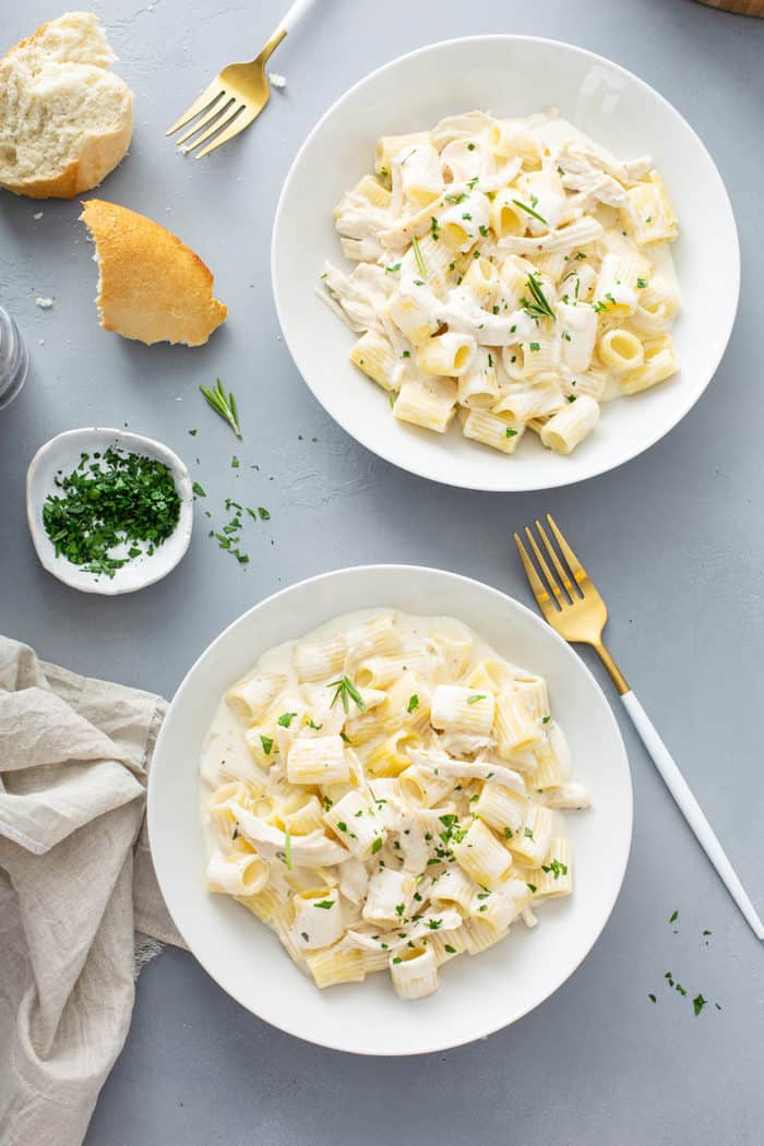 Two white bowls of goat cheese pasta on a gray countertop next to chopped herbs and baguette pieces