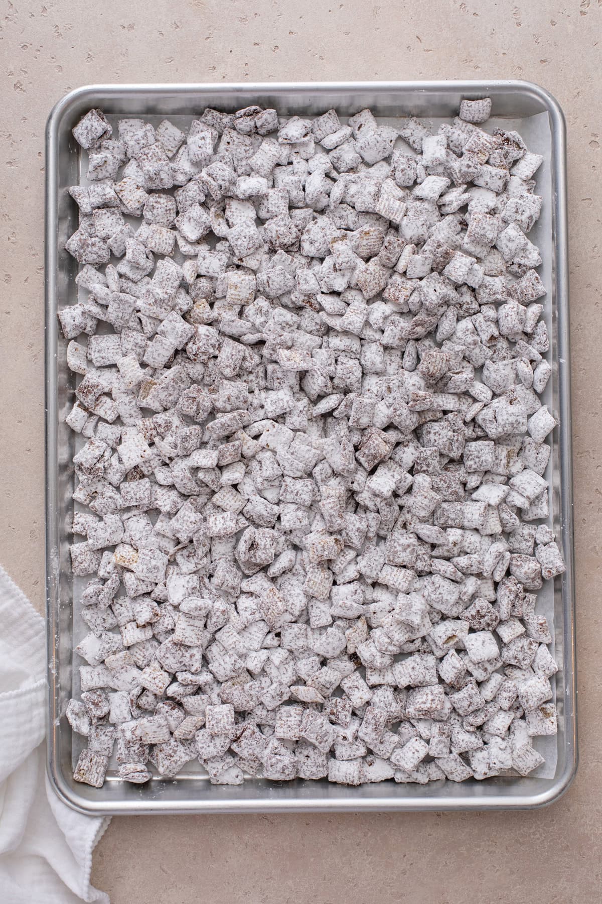 Puppy chow coated in powdered sugar and cooling on a rimmed baking sheet.