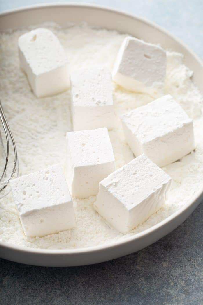 Homemade marshmallows being dredged in cornstarch and powdered sugar in a shallow white bowl