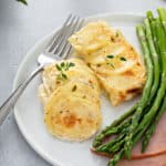 Close up of scalloped potatoes next to a fork, asparagus and ham on a white plate