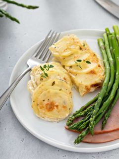 Close up of scalloped potatoes next to a fork, asparagus and ham on a white plate