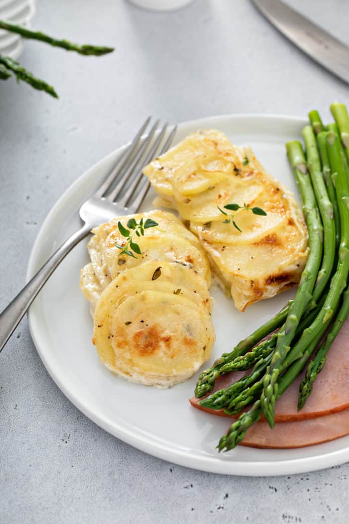 Close up of scalloped potatoes next to a fork, asparagus and ham on a white plate