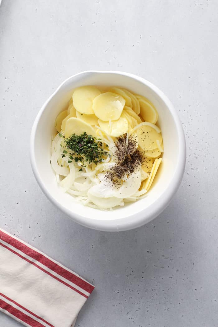 Sliced potatoes, onions, and seasonings in a white mixing bowl on a gray countertop