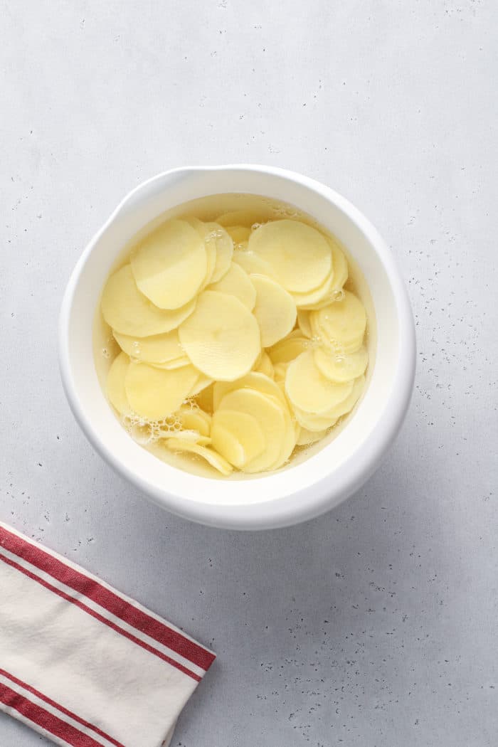 Sliced potatoes soaking in water in a white mixing bowl