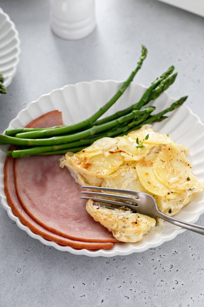 Fork about to take a bite of scalloped potatoes that are on a white plate next to ham and asparagus