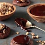 Small bowl of melted chocolate and a bowl of crushed Heath bar next to Heath cookies and a spoon on a stone surface