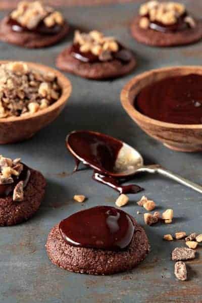 Small bowl of melted chocolate and a bowl of crushed Heath bar next to Heath cookies and a spoon on a stone surface