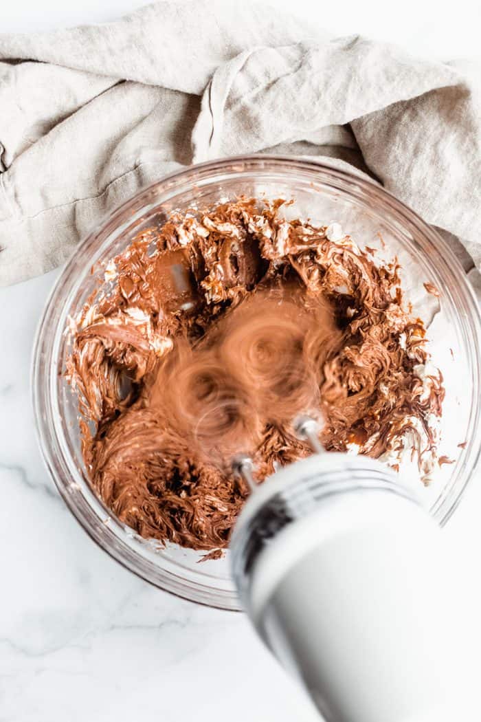 Overhead view of electric mixer beating together nutella and cream cheese in a glass mixing bowl for no bake nutella cheesecakes