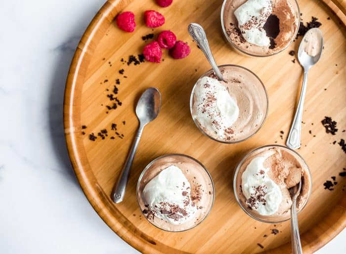 Overhead view of four mini no bake nutella cheesecakes on a wooden board, surrounded by spoons, chocolate shaving and raspberries