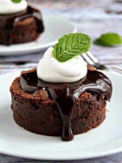 A mint brownie on a round plate topped with hot fudge, whipped cream, and a fresh mint leaf