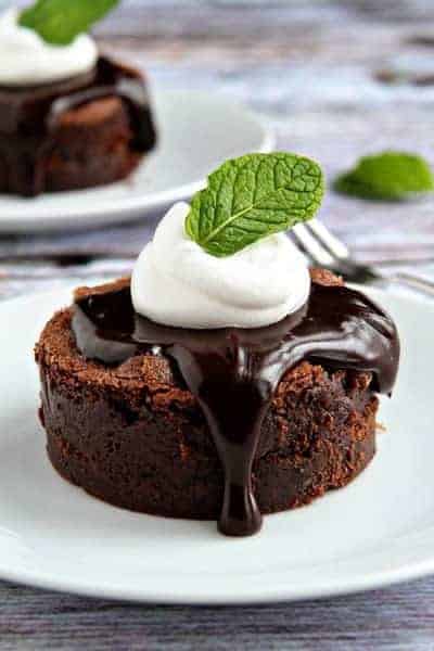 A mint brownie on a round plate topped with hot fudge, whipped cream, and a fresh mint leaf